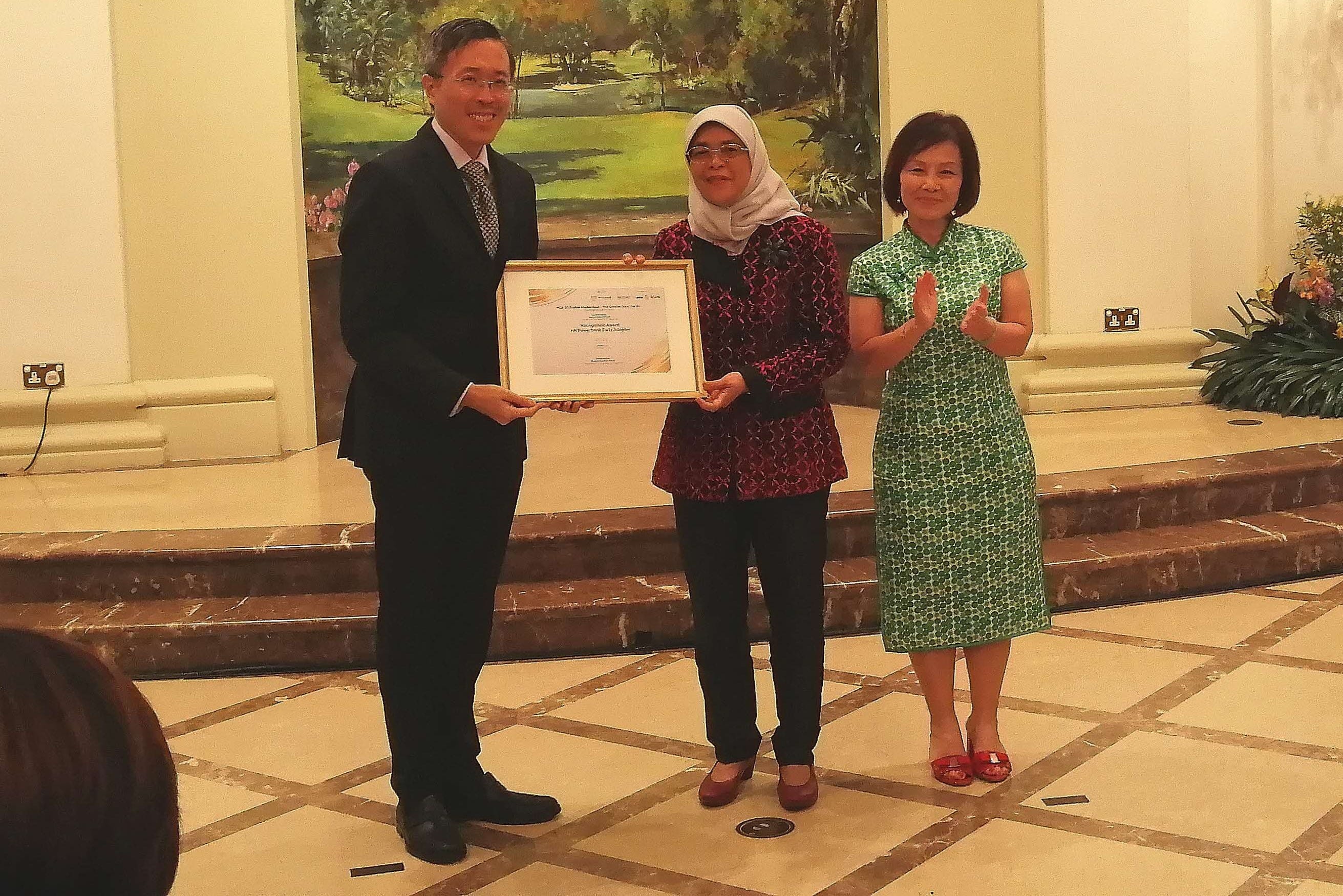 Two people hold a framed certificate while a third woman in green applauds beside them indoors.
